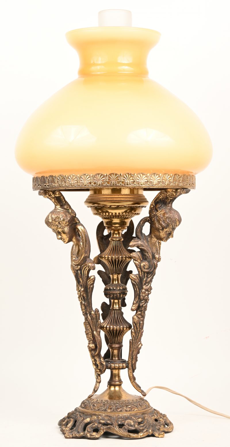 Een vintage tafellamp in kunstbrons. Gesimuleerde olielamp met geel wit glazen lampenkap. 3 poten en center met gesculpteerd reliëf en dameshoofden afgebeeld.