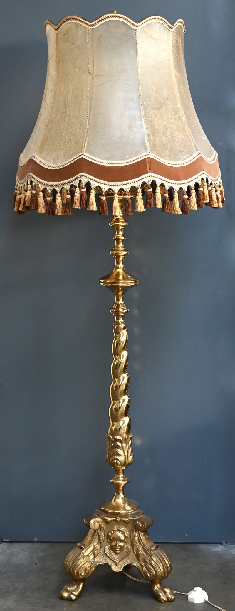 Een grote messing lampadaire met diverse ornamenten en staande op drie poten. Organische elementen en gezichten in het reliëf. Lampenkap uit vermoedelijk varkenshuid met flosjes onderaan.