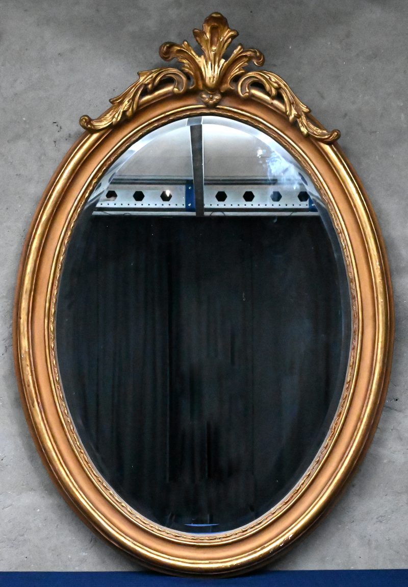 Een grote, ovale spiegel verguld en met ornament bovenaan.