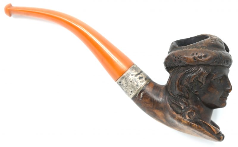 Een pijp met pijpenkop van gebeeldhouwd hout in de vorm van een Schillerachtige figuur. Metalen aansluiting aan amberkleurige steel.