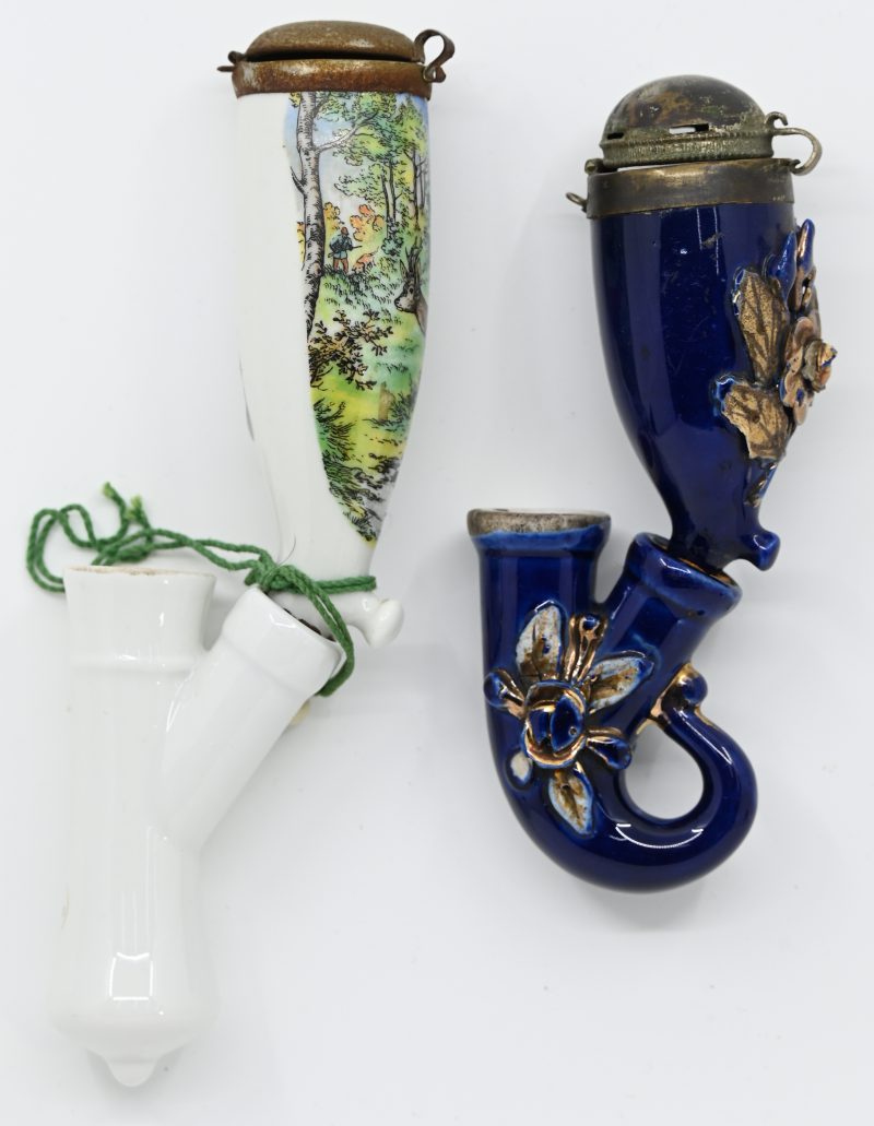 Twee porseleinen pijpenkoppen met vochtsluis. De éne met een decor van een hertenjacht, de andere kobaltblauw met een vergulde bloementuil in reliëf. Beide met metalen deksel.