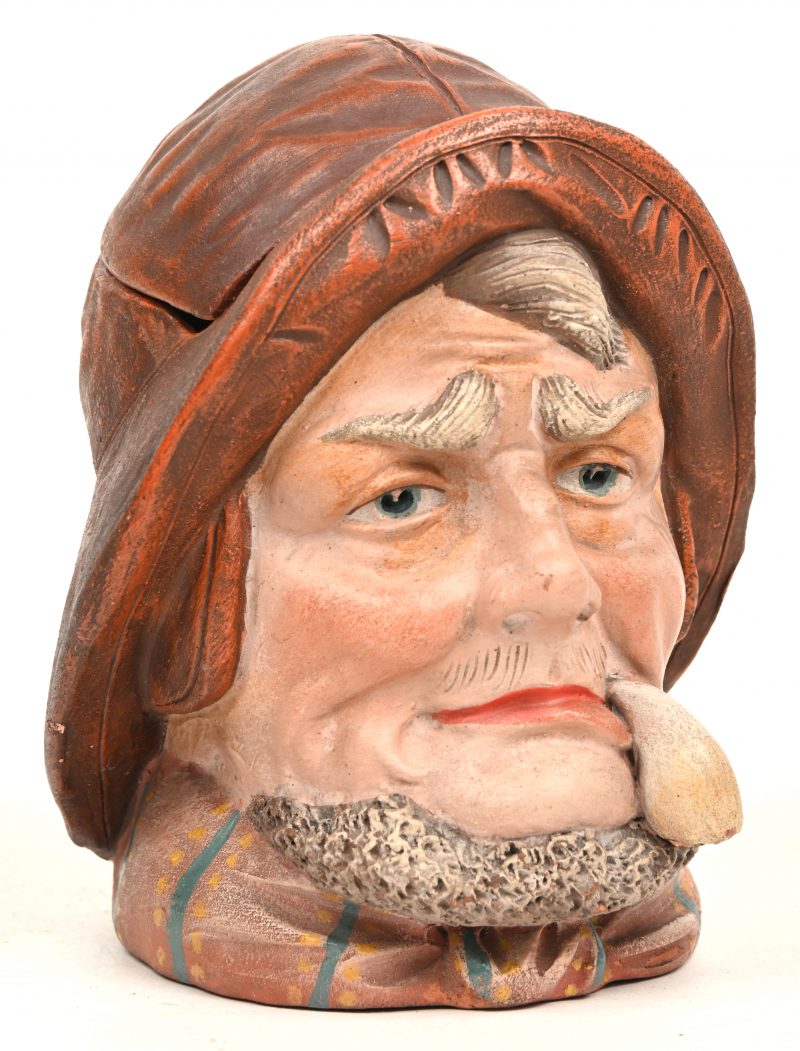 Een tabakspotje van polychroom steengoed in de vorm van de kop van een pijprokende zeeman. Onderaan gemerkt G. Hanns (?).