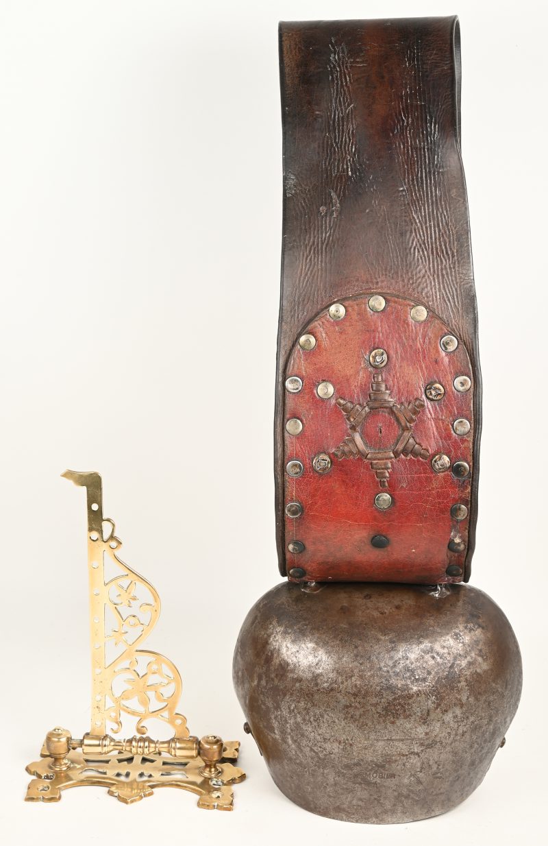 Een grote Zwitserse koebel met diklederen beslagen, halsband. Gemerkt “P.Morier” N.4. Vervaardigd in Morges, Zwitserland. Bijgevoegd een muurhouder.