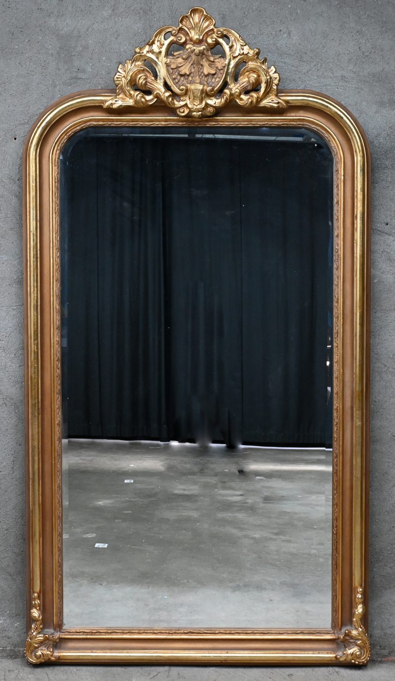 Een groot formaat spiegel met verguld houten kader, ornament bovenaan en geslepen glas.