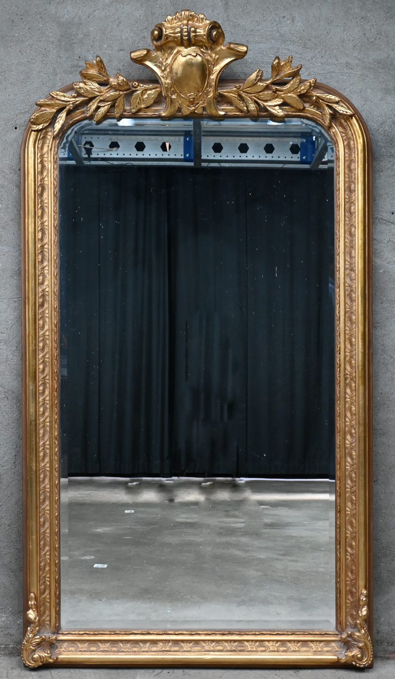 Een groot formaat spiegel met verguld houten kader, ornament bovenaan en geslepen glas.