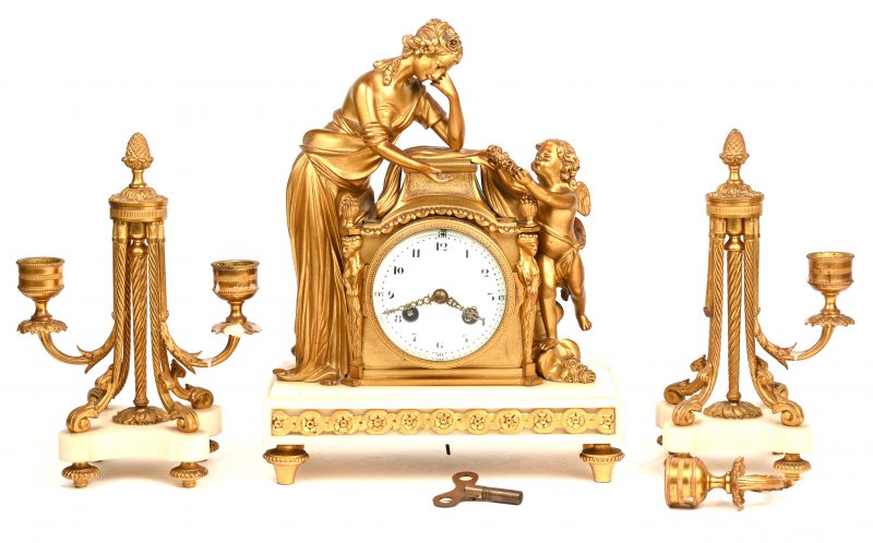 Een Charles X-stijl klokkenstel van verguld brons en albast. De pendule versierd met een jonge vrouw, die door een engel een bloemenkrans wordt aangeboden. Vergezeld van twee kandelaars met twee lichtpunten (één arm om opnieuw te bevestigen).