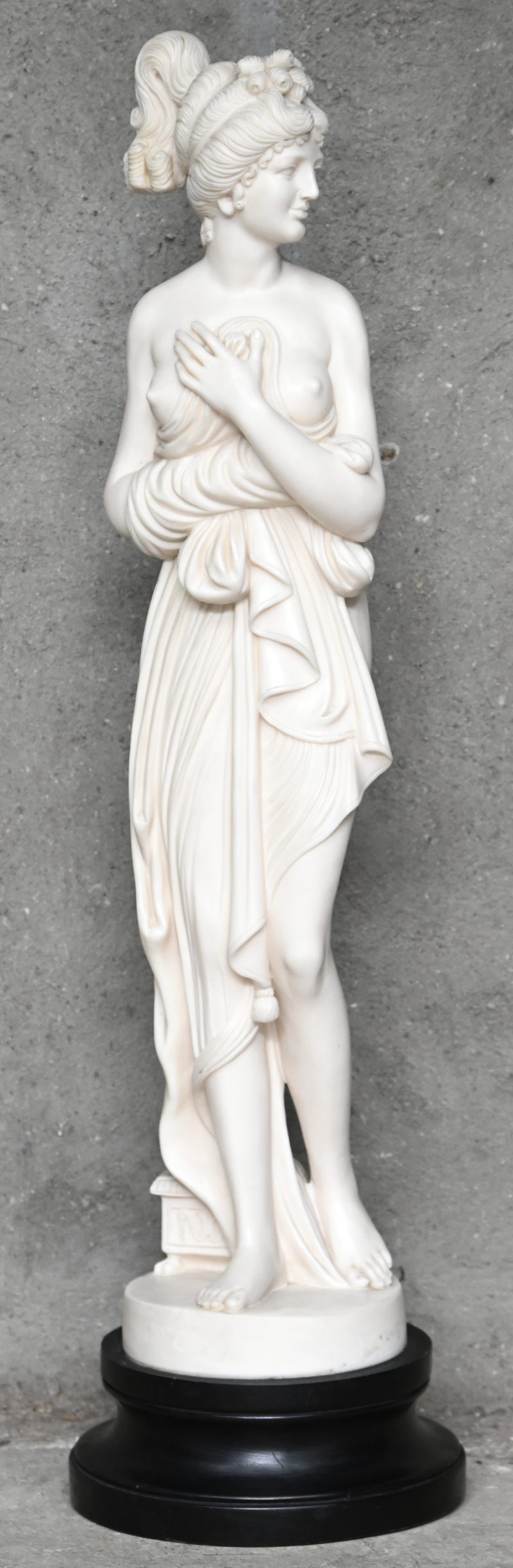 Een uit kunststof gesculpteerd beeld van een gesluierde Venus Italica met ontblote borst, op houten voet.