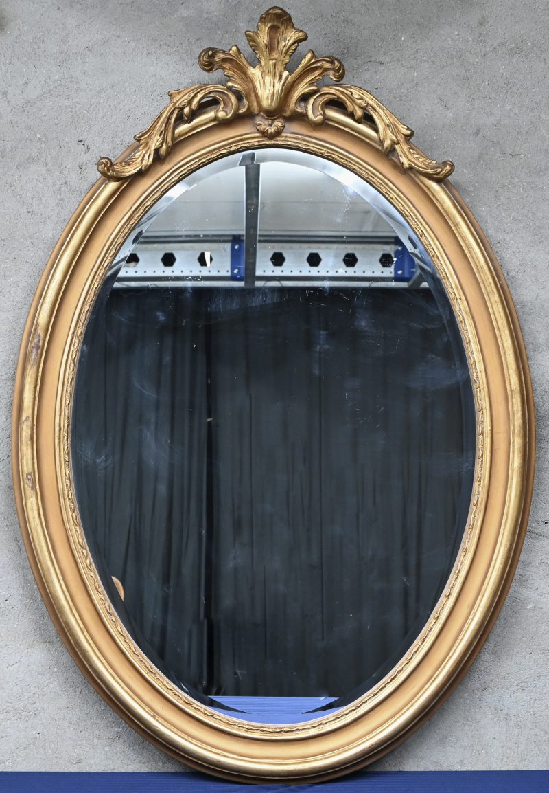 Een ovale spiegel met verguld kader en gesculpteerd ornament bovenaan.