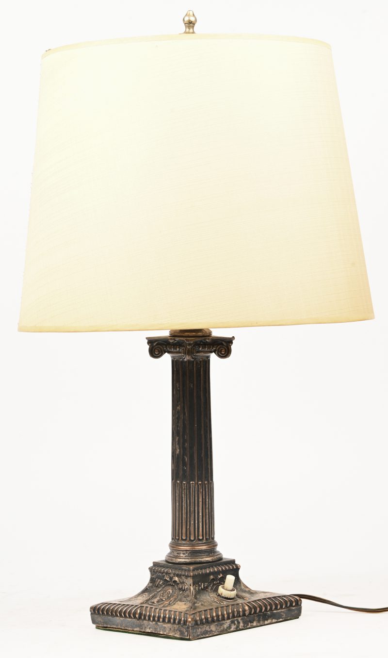 Een tafellamp in de vorm van een zuil met onderaan de afbeelding van een Romein en guirlandes. Deze is ooit verzilverd geweest.
