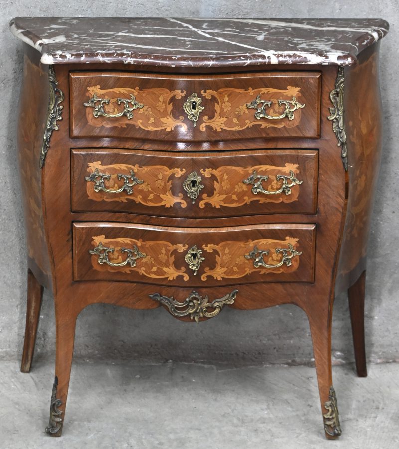 Een kleine commode in Lodewijk XV stijl. Dubbel gebogen front met drie laden, marmeren blad, gemarquetteerde versiering.