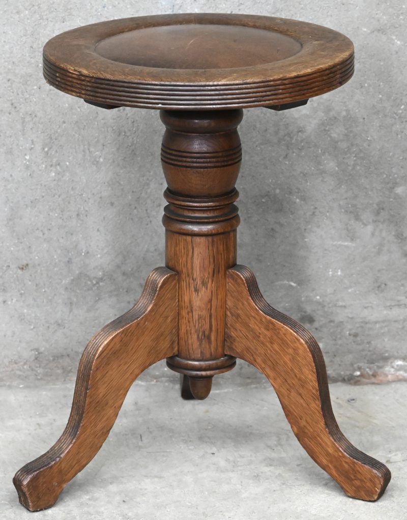 Een houten pianstoel met rond tablet en lederen zitting. Verstelbaar in hoogte met schroef, gesteund op drie poten. Onderaan genummerd 4053.