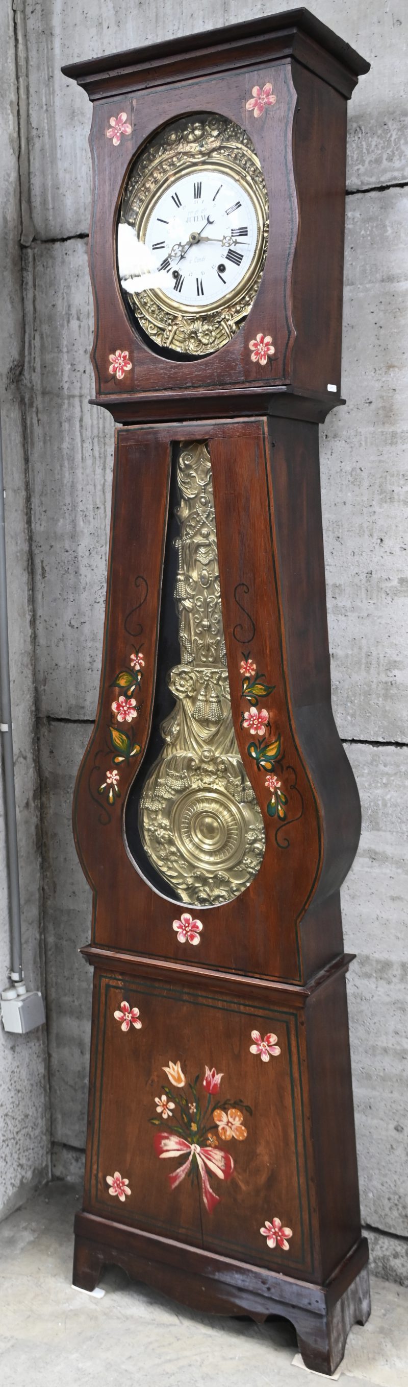 Een Franse comtoise klok met opschrift op de wijzerplaat Juteau à Candé in een houten kast met geschilderde bloemen versierd.