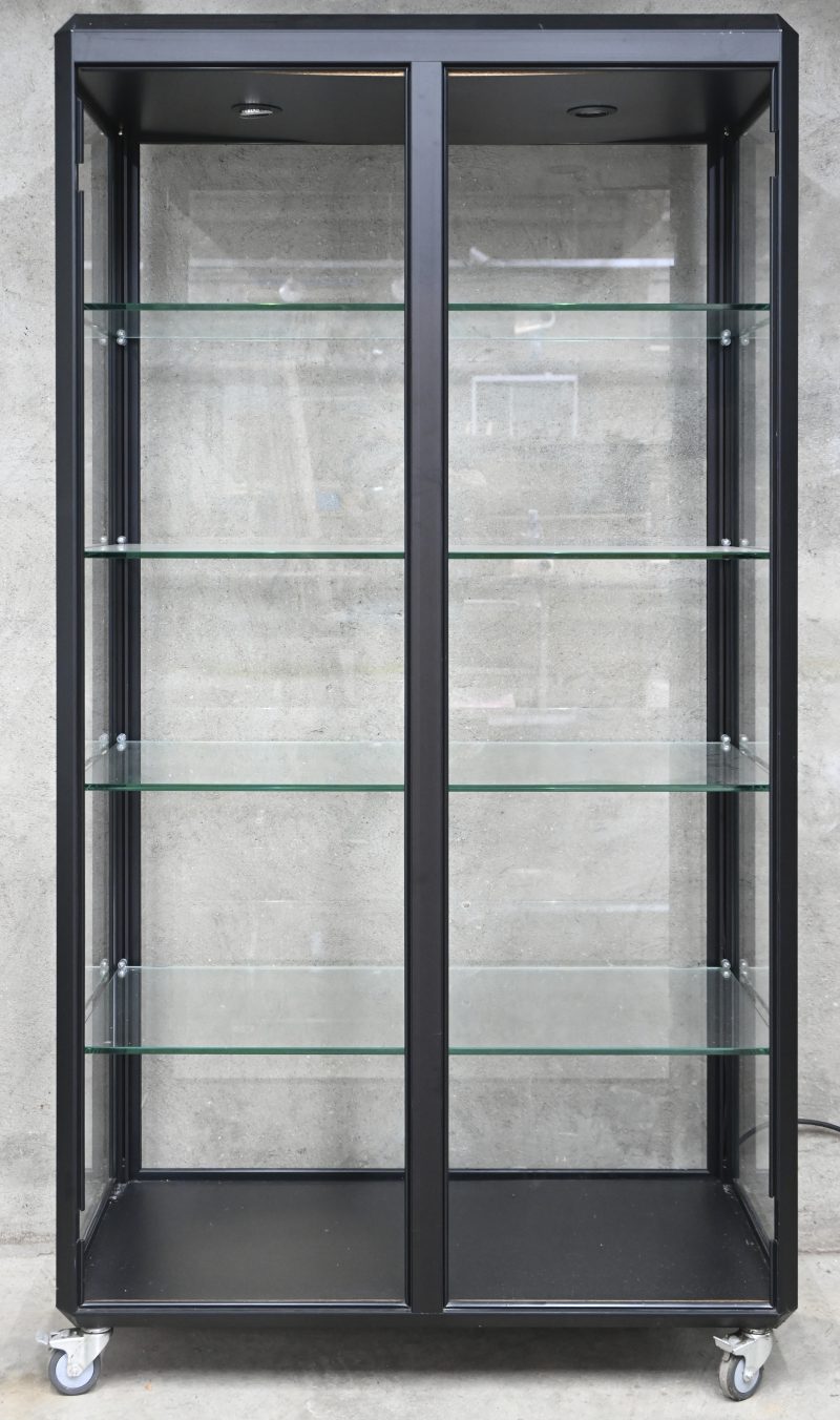 Een glazen vitrine op wieltjes met zwart frame. 4 schabben.