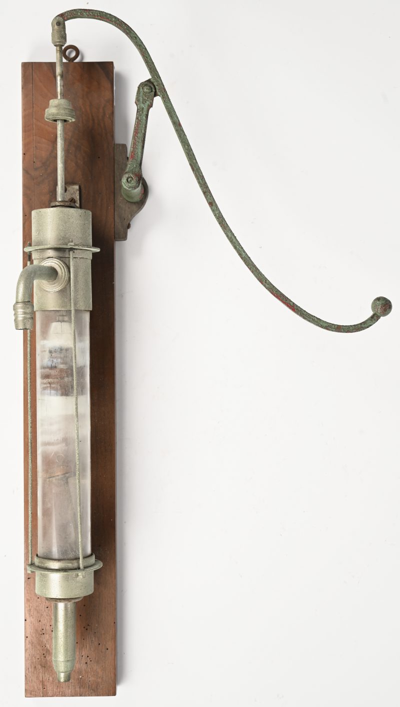 Een oud model pomp in metaal en glas aan een houten plank voor wandbevestiging.