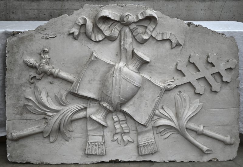 Een wit plaasteren bas-reliëf met een embleem bestaande uit een boek, scepter, urn enz...