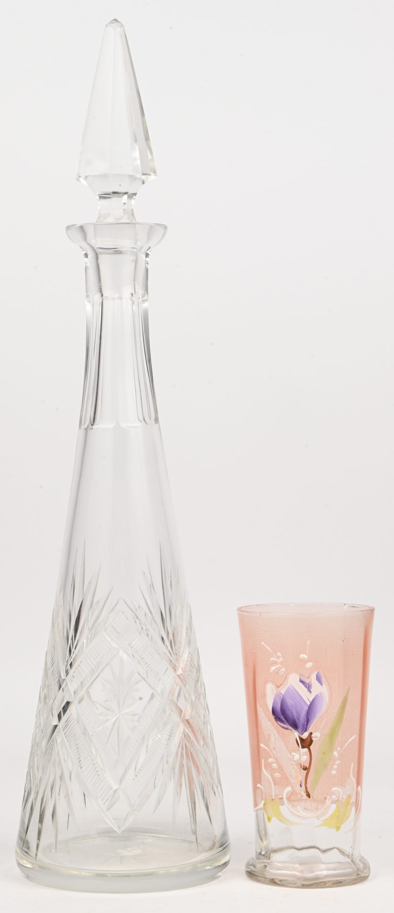 Een kleurloze, geslepen glazen kegelvormige karaf, evenas een roze glazen glas versierd met een koudgeschilderde krokus. Omstreeks 1900.