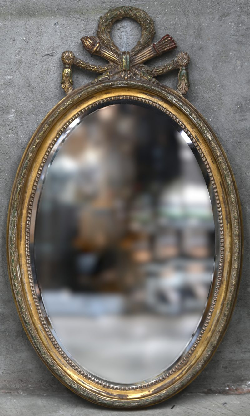 Een ovale spiegel in verguld kader met polychrome restanten en gesculpteerde details boven. Begin 20e eeuw.