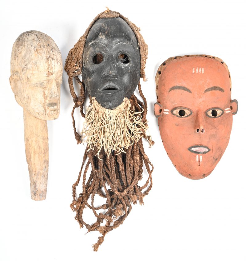 Een lot van drie Afrikaanse maskers in hout waarvan één rood, één zwart met witte baard en één wit hoofd met handvat.