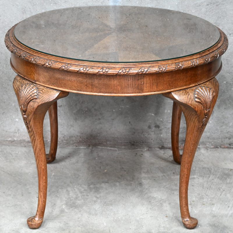 Een rond houten salontafel met gesculpteerde details en geometrisch ingelegd patroon. Bovenop een glazen blad.