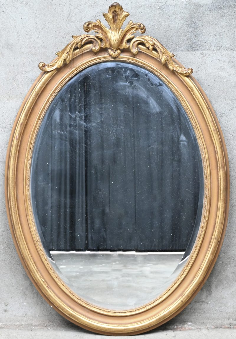 Een verguld ovalen spiegel met gesculpteerd ornament bovenaan het kader.