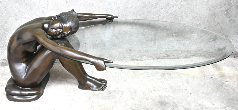 Een bronzen salontafel van een zittend, naakte vrouw, dewelke een glazen ovaal tafelblad vasthoudt.