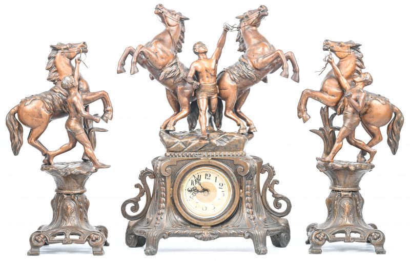 Een schouwgarnituur van zamak met een klokje en twee beeldjes op een sokkeltje. Met de voorstelling van paarden en paardenmenners naar Coustou.
