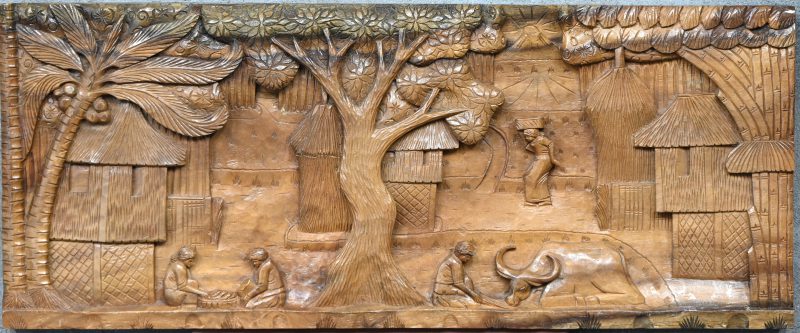 Een Aziatisch houtsculptuur met de voorstelling van een dorp, enkele figuren en palmbomen.