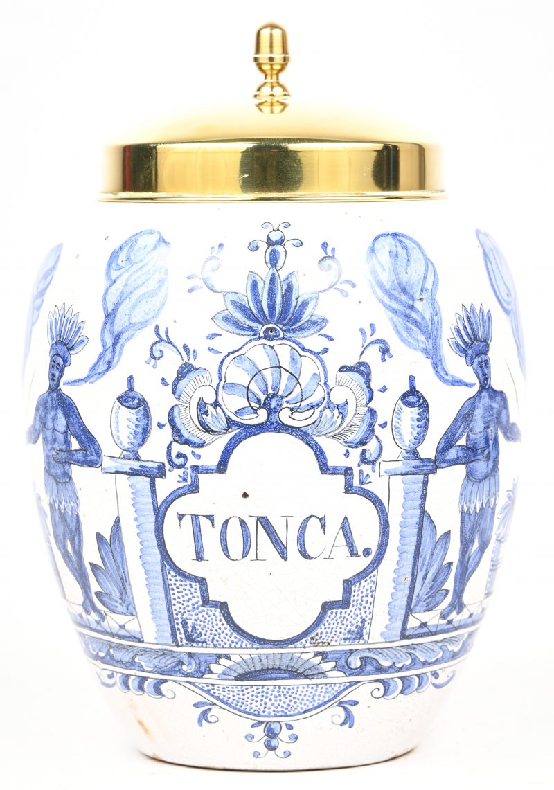 Een Delfts blauw-wit aarderkwerken tabakspot met messing deksel. Decor met rokende Indiaan en opschrift “TONCA”. Onderaan gemerkt “De Porceleyne Claeuw”.