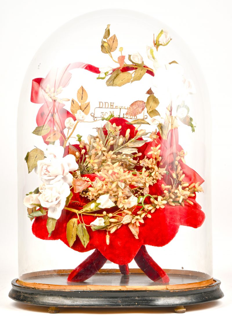 Een antieke bloemenkrans met porseleinen en kunstoffen bloemen, onder glazen stolp. Tekst binnenin; “DDH unisa EV 30 M. 1906”