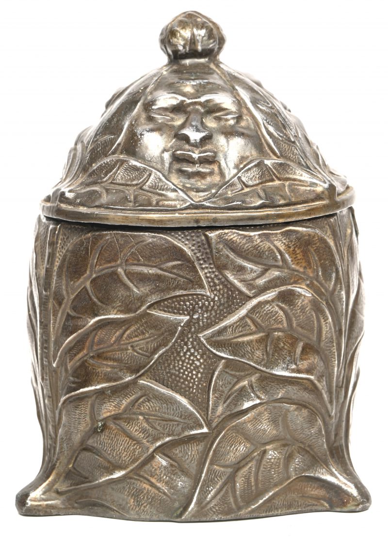 Een ronde theedoos van brons met een vegetaal reliëfdecor met op het deksel drie gezichten. Klein letsel aan de bovenrand.