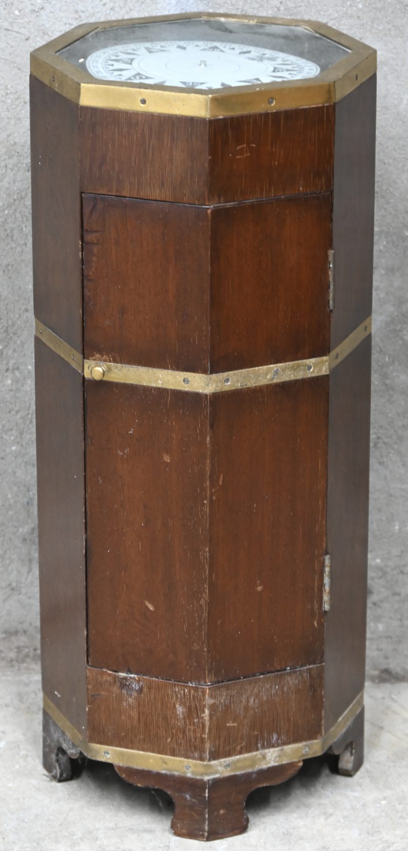 Een kompas onder glasplaat in 8-hoekig houten kastje met 1 deur en messing omlijstingen.