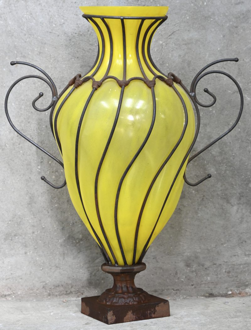 Een grote vaas, geel glas in een smeedijzeren omhulsel op gietijzeren voet.