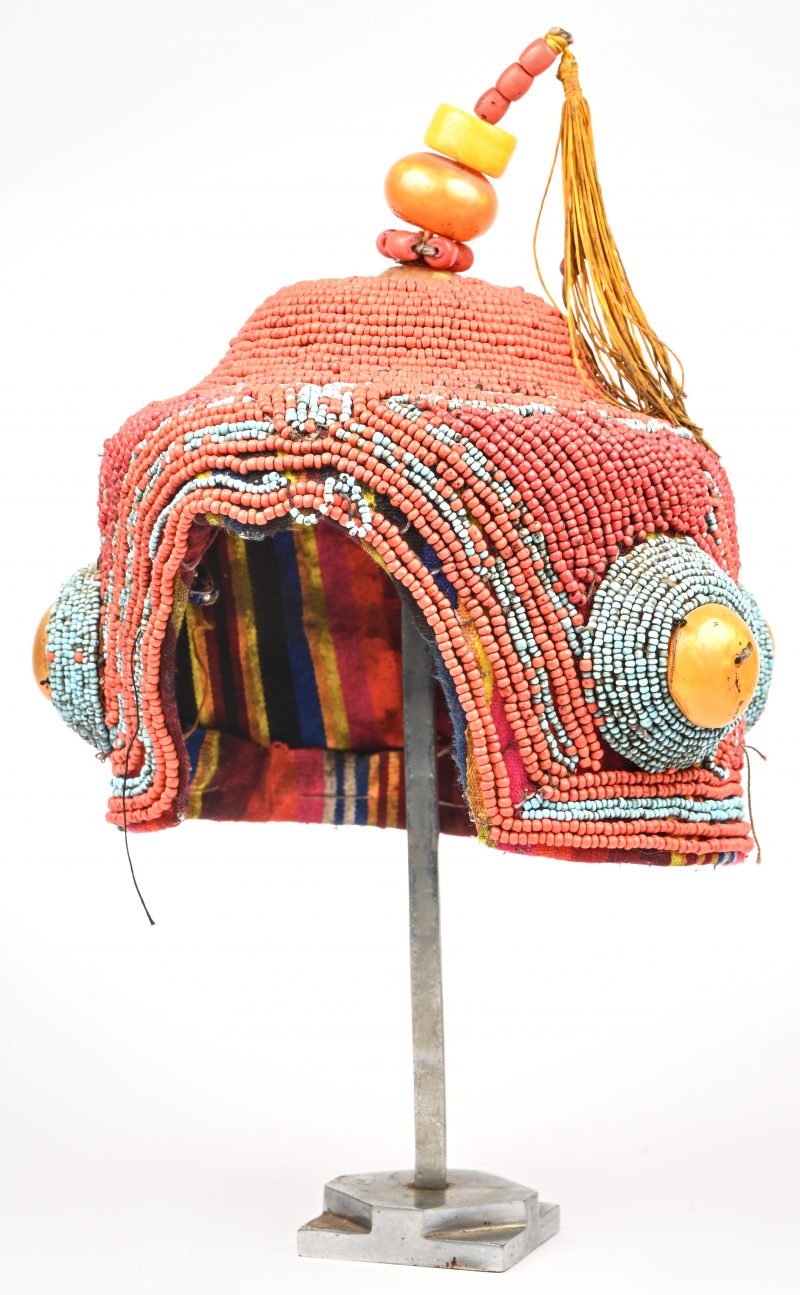 Een meerkleurig Tibetaans of Chinese, helmvormige trouwhoed. Versierd met diverse kralen, cabochons uit kopal en gestoffeerde voering. Op metalen staander.