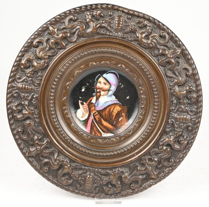 Een ronde plaquette van veelkleurig porselein meet voorstelling van een Spaanse soldaat. In een ruime omlijsting van gedreven koper.