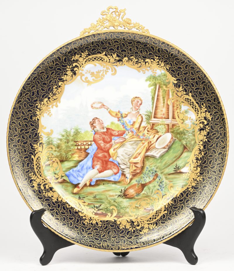 Een porseleinen sierbord met romantisch figuratief decor en vermelding “R. Charlotte”, vergulde rand en ornament. Verso gemerkt.