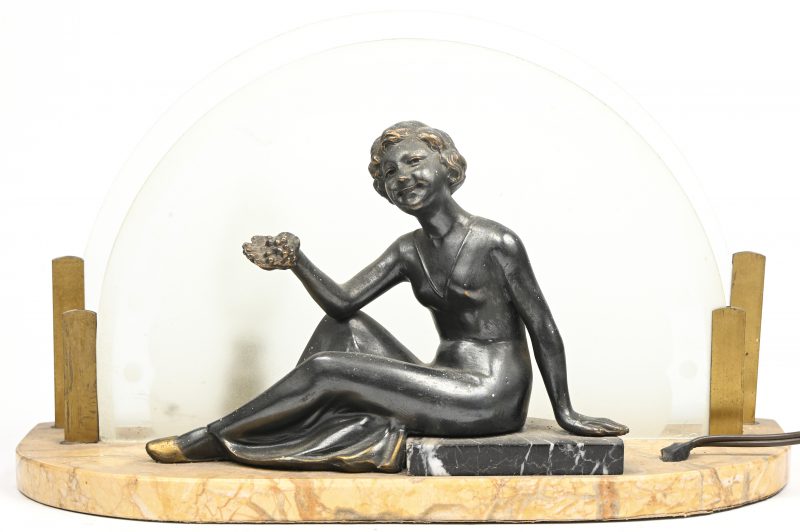 Een art-deco tafellamp met een bronzen beeld van een druivenmeisje gezeten op marmer. Achteraan een lamp tussen twee halfronde glazen platen.