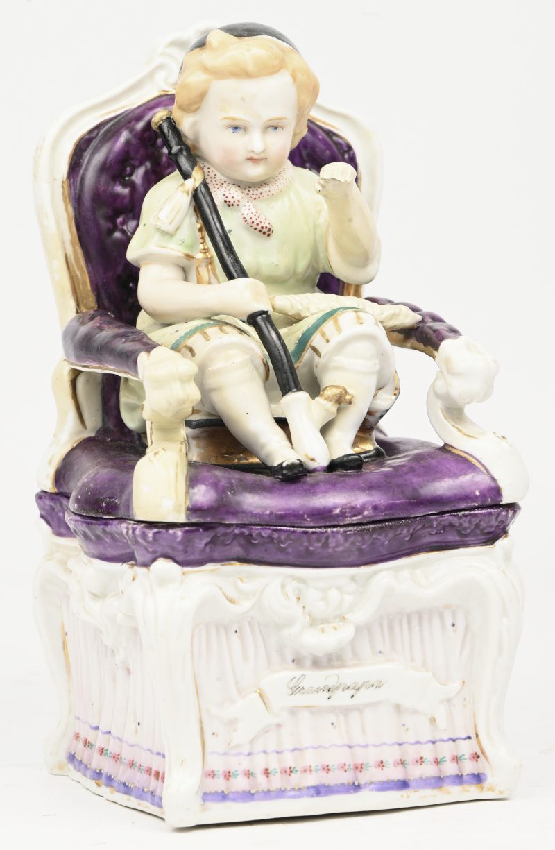 Een polychroom porseleinen beeldje van een kindje op een stoel als tabaksdoos. Opschrift vooraan “Grandpapa”. Boekje in hand afgebroken.