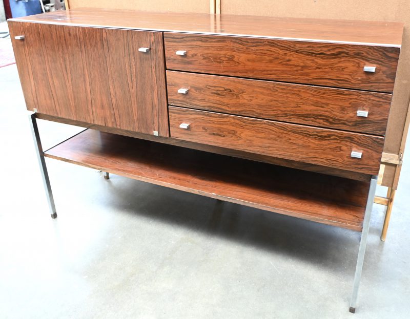 Een midcentury sideboard/barmeubel in pallisanderfineer op chrome poten. Eén deur en drie laden. Kleine schade aan de hoek links achter.