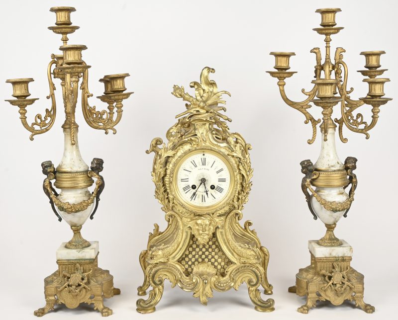 Bronzen mantelklok in barokke stijl met vergulde delen, slinger en glas manko. Toegevoegd een paar 6 armige kandelaars, waarvan telkens één arm manko.