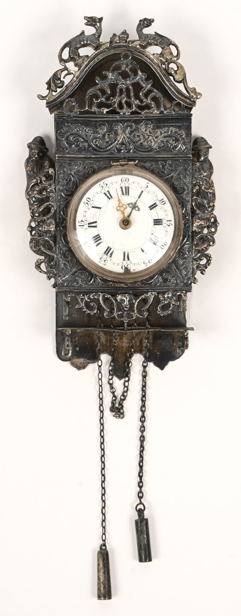 Een horlogehouder van zilver met Hollandse keuren (tweede gehalte), in de vorm van een wandpendule met filigraanwerk. Voorzien van een zakhorloge met messingen kast en met wijzerplaat van wit email. XIXde eeuw.