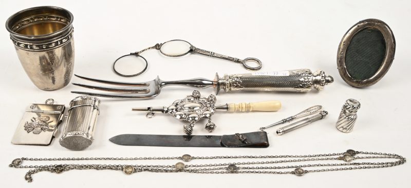 Een gevarieerd lot zilveren items met een variatie aan keuren bestaande uit een beker, een notaboekje, een halsketting, een luciferdoosje, een parfumflesje, een brievenklem, een haakje, een brilletje, een serveervork, een kadertje, een potlood en een rammelaar. 195 gram.