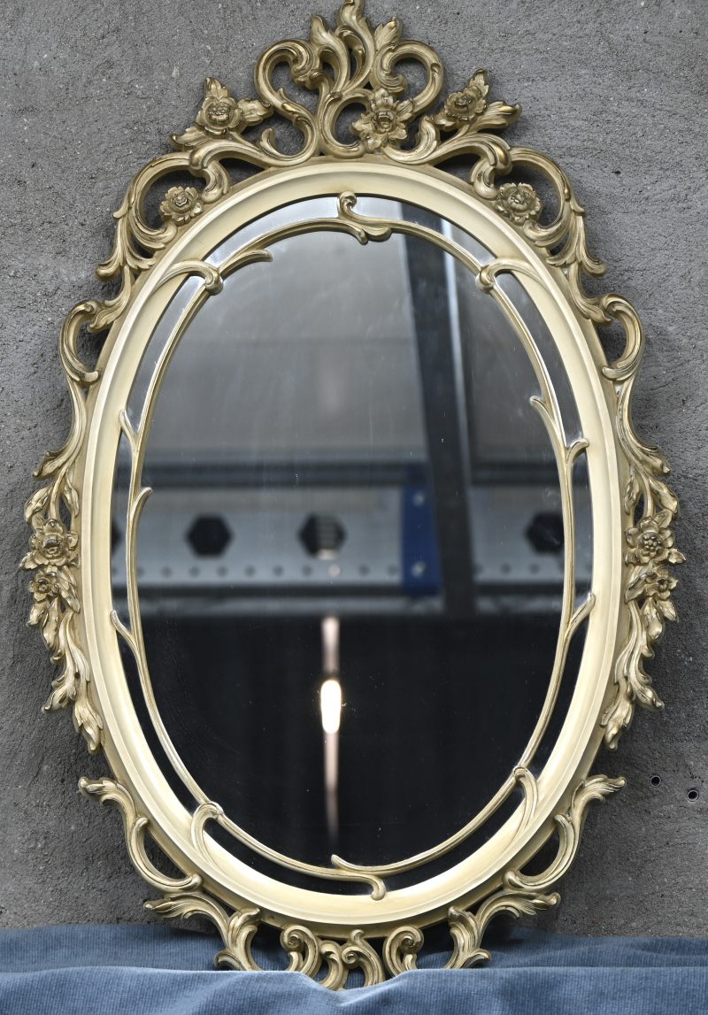 Een ovale spiegel met barokke lijst van crèmekleurig hout.