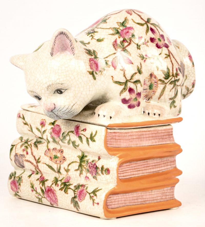 Een porseleinen bonbonnière voorstellend een poes op 3 boeken, onderaan een merkteken AN en een datum naar welke periode het gemaakt is.