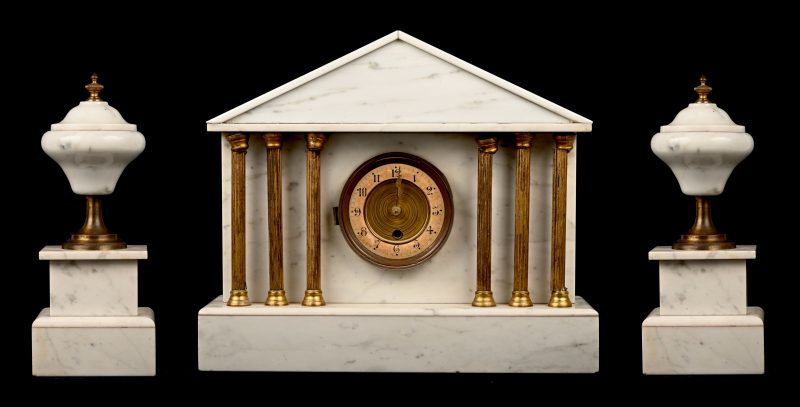 Een witmarmeren klokstel in de vorm van een Griekse tempel met Dorische zuilen. Met bijbehorende urnvormige ornamenten. De klok is te herstellen.