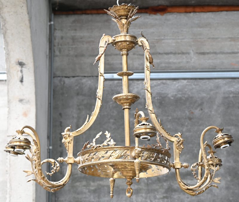 Een bronzen luchter met vier armen en zes lichtpunten, twee lampen in de basis. Gedecoreerd met ramskoppen en lauwerkransen. Zonder blakers.