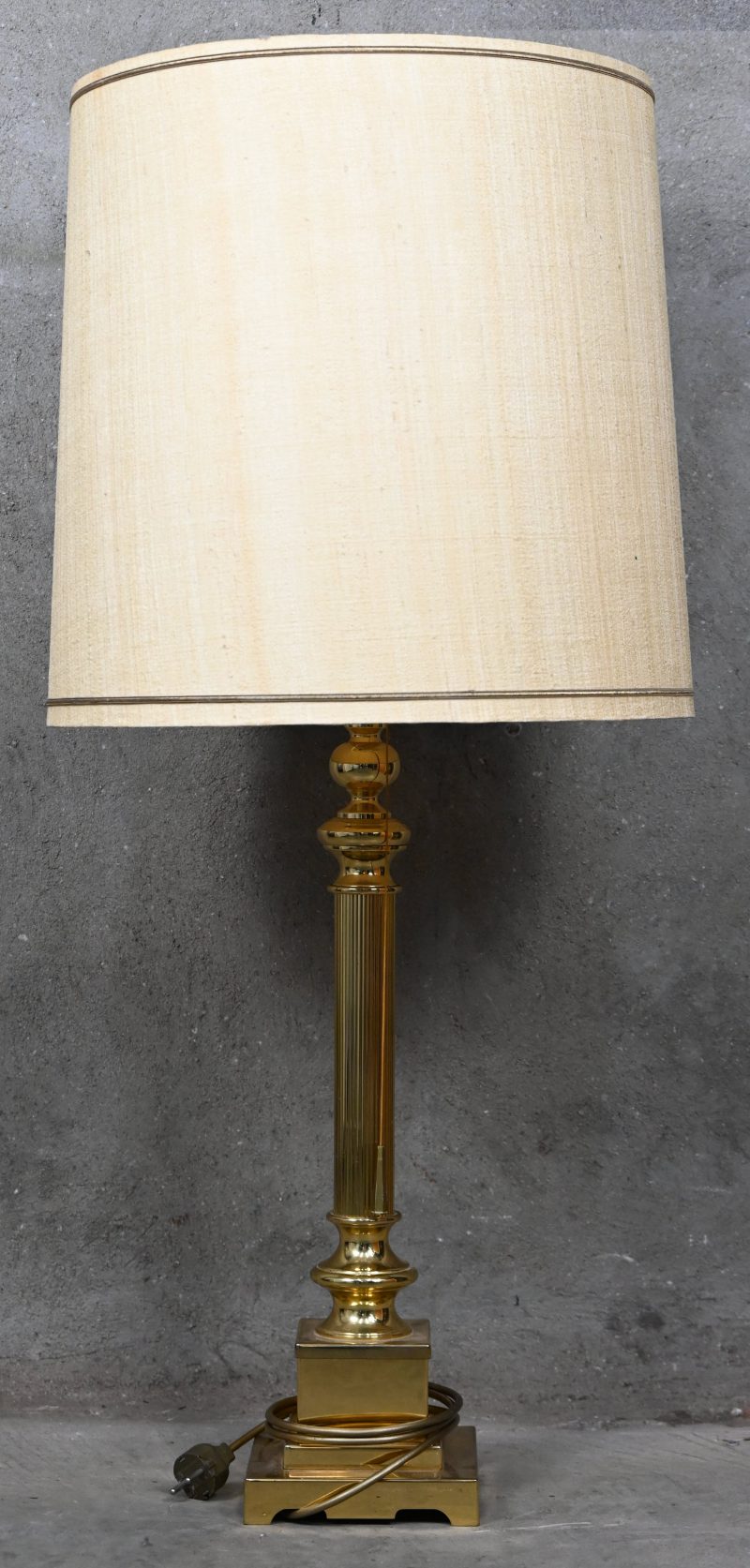 Een grote tafellamp op goudkleurige zuil met een stoffen ecrukleurige kap. Twee lichtpunten.