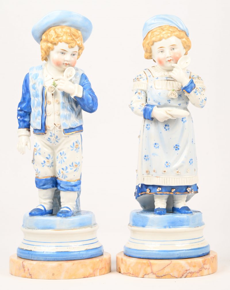 Een paar polychrome porseleinen beeldjes van een jongen en een meisje, op marmeren voet.