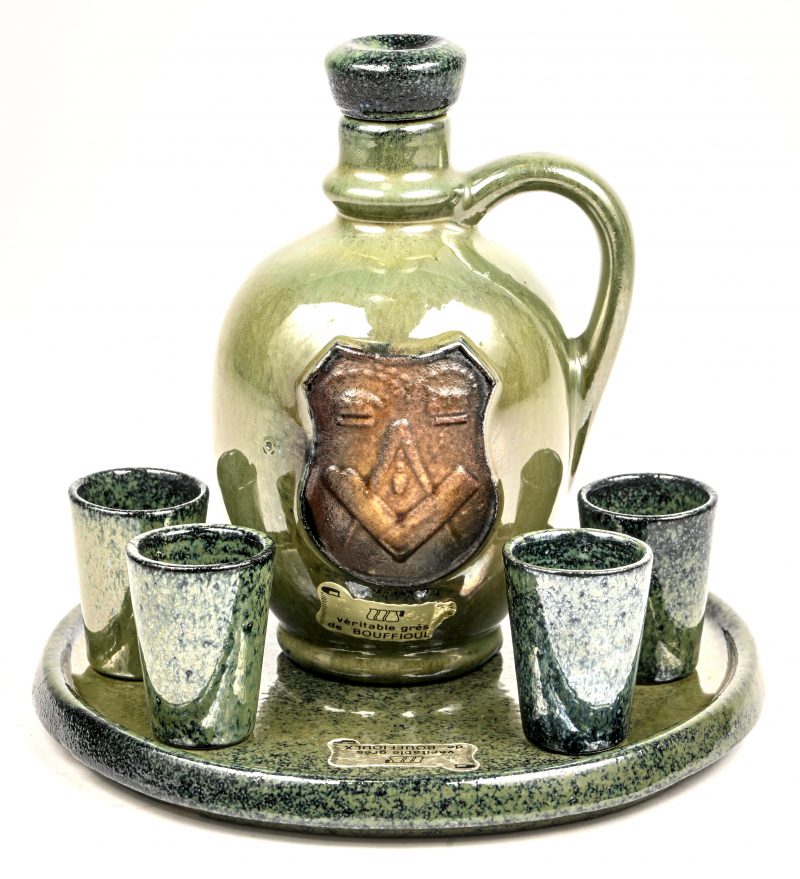 Een karaf met borreltjes op dienblad van gres aardewerk. De kruik draagt het embleem van de vrijmetselaarsloge. De kurk van de stop is manco.
