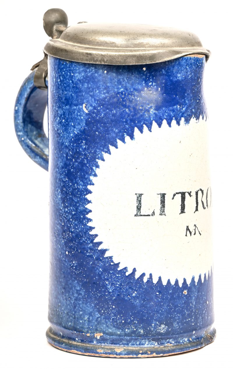 Een antieke schenkkan met deksel. Van blauw geglazuurd aardewerk met opschrift; Litron. Er loopt een barst over de gehele hoogte van de kan, hersteld met metalen krammen.