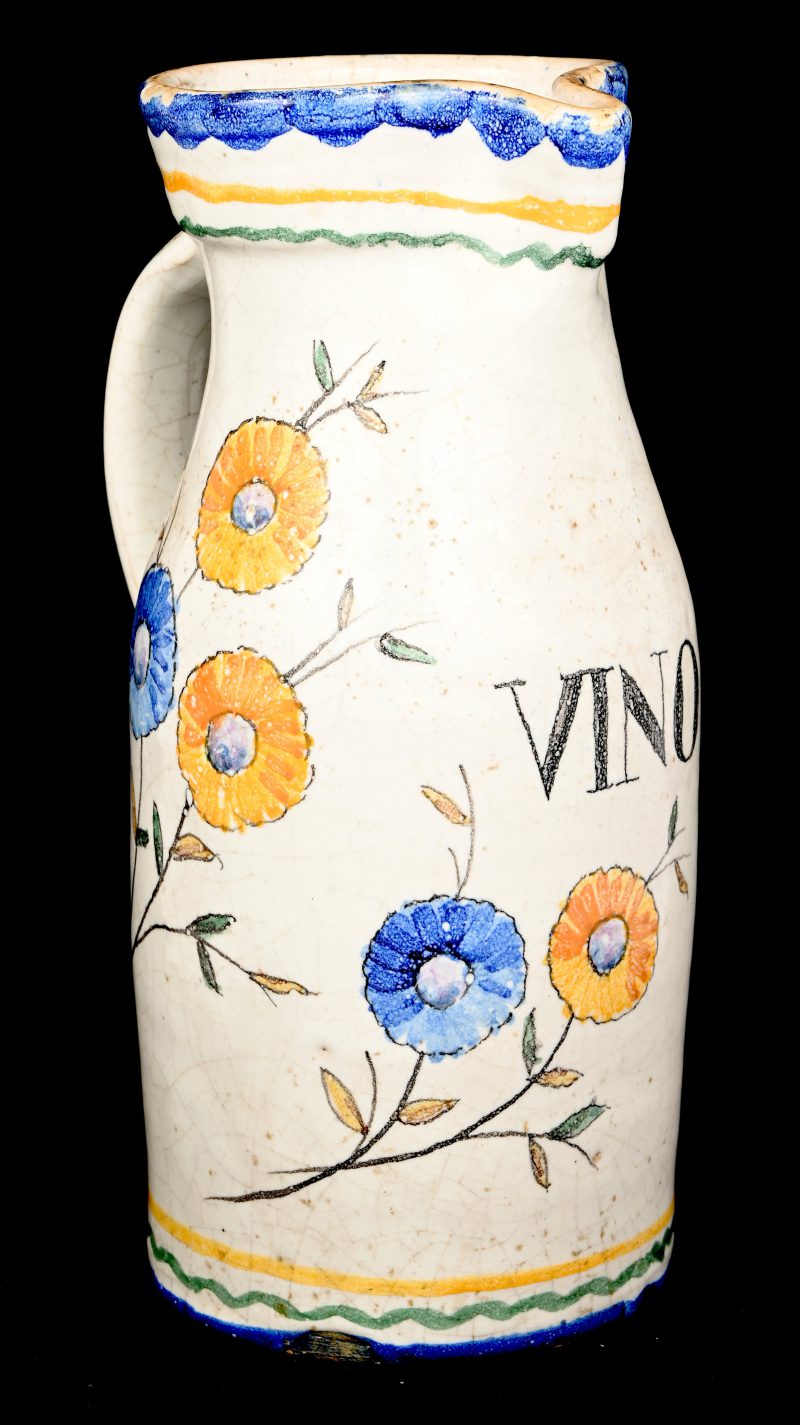 Een antieke schenkkan van geglazuurd aardewerk. Met een decor van bloemen en het opschrift; VINO.
